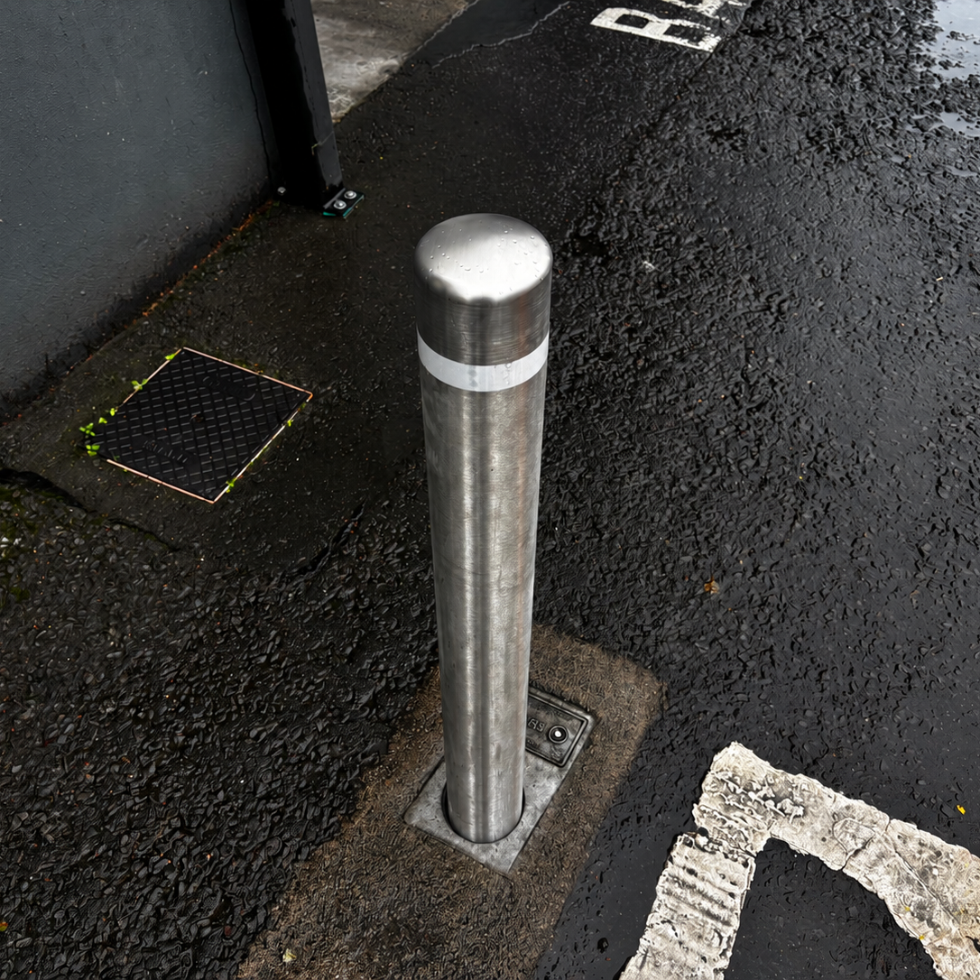 A stainless steel bollard post with a reflective silver band, installed on a wet asphalt street corner next to a dark gray wall.