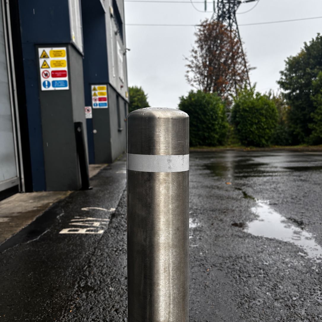 A stainless steel bollard post with a reflective silver band, situated outdoors on a wet asphalt surface next to a dark blue building.