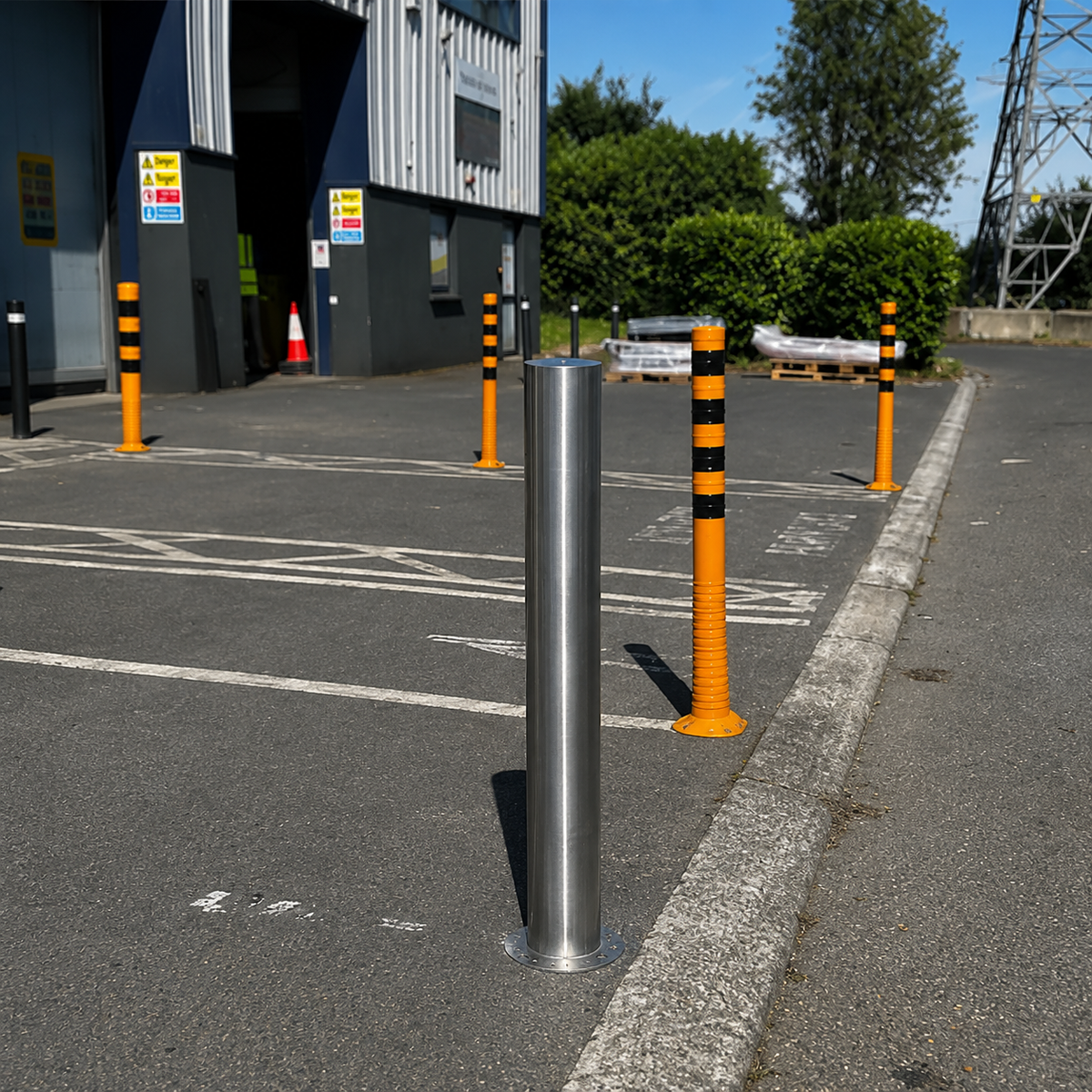 A stainless steel cylindrical bollard post installed in an outdoor paved area next to a building, with several orange and black striped posts around it.