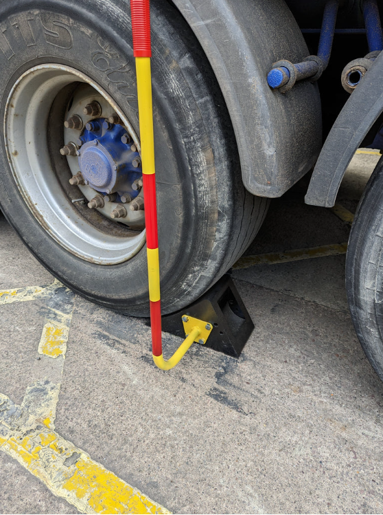 A Handled Wheel Chock placed on the ground next to a truck wheel, with a steel handle and yellow reflective strips on it.
