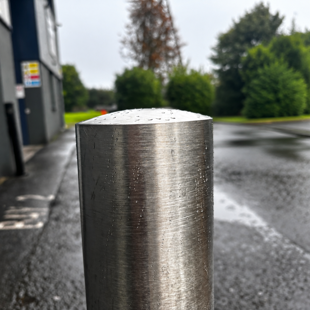 A close-up of a cylindrical brushed metal bollard post on a wet asphalt surface, with a building and trees blurred in the background.