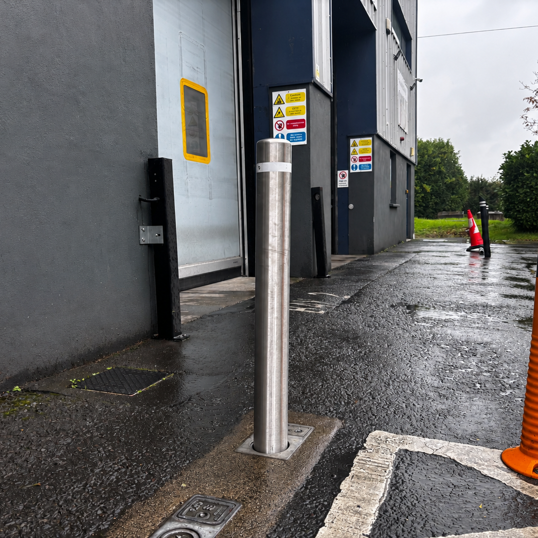 Outdoor industrial scene showing two bollards: a tall silver metal post set on a wet paved area beside a gray building.