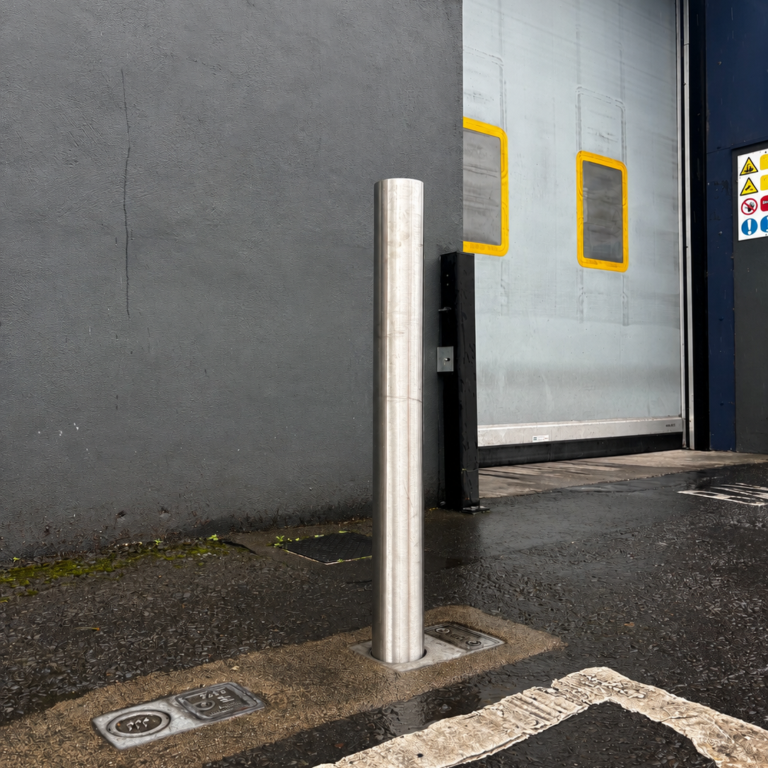 A tall, cylindrical stainless steel bollard post installed on a wet asphalt driveway in front of a gray industrial wall and a large white garage door.