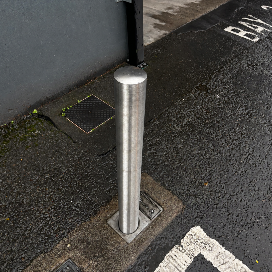 A cylindrical stainless steel bollard post installed on a wet asphalt street corner next to a dark gray wall, with painted white road markings and a drain visible.